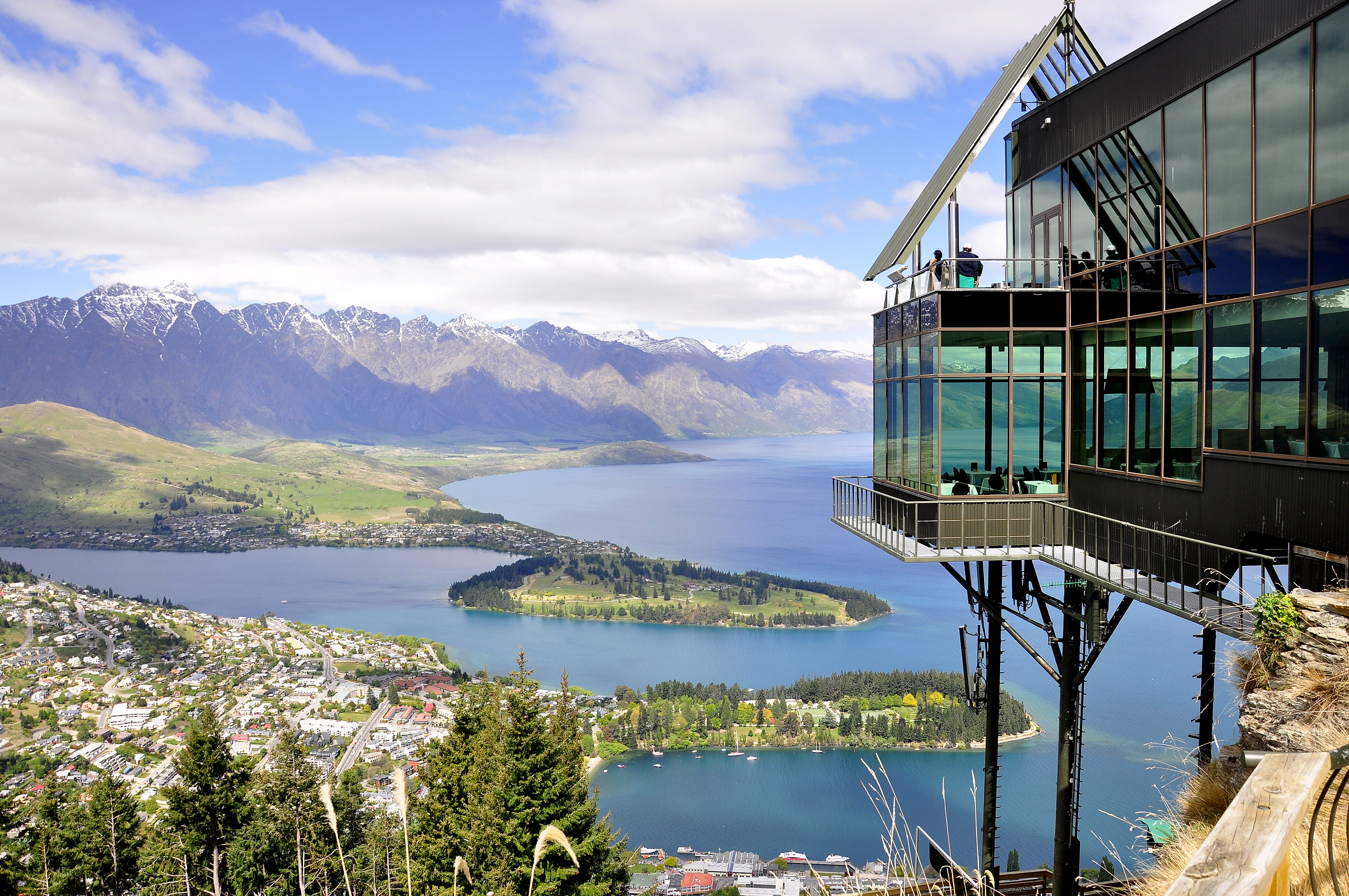A large glass building sits on a high hill overlooking the Queenstown skyline of snow-capped mountains and a large lake with a chunk of greenery jutting into the water
